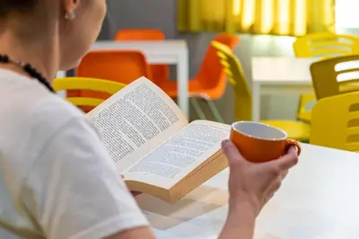 a woman sitting at a table with a book and a cup of coffee at Green Cube Capsule Hostel in Sofia