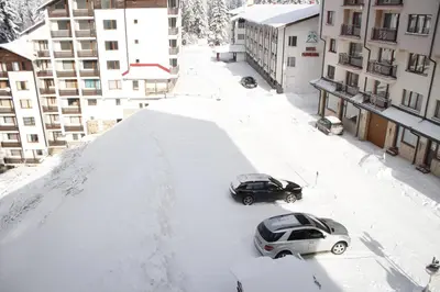 two cars parked on a snow covered street with buildings at Nevada Aparthotel in Pamporovo