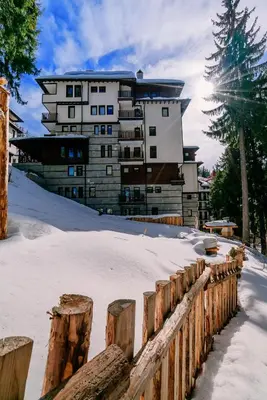 a building in the background with snow and a fence at Green Life Family Apartments Pamporovo in Pamporovo
