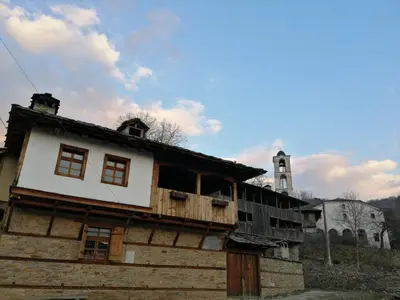 an old wooden building with a church in the background at Къща за гости Кануш войвода in Kovachevitsa