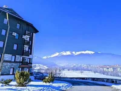 a building in the snow with mountains in the background at Stylish 2BD Apartment with Panoramic Mountain View by Flat Mаnager in Bansko